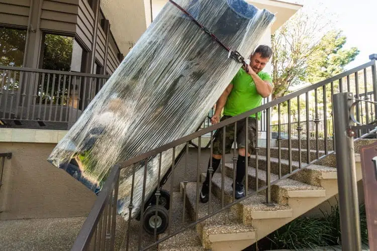 A person in a green shirt moves a large, wrapped object on a dolly down an outdoor staircase, perhaps part of their journey moving from Los Angeles to Miami.