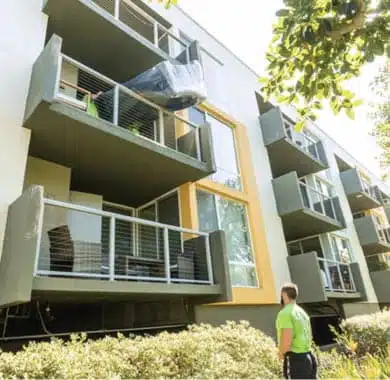 A person in a green shirt stands outside an apartment building, looking up at a balcony where a large object is being moved down using a rope, likely part of their move from Los Angeles to Miami.